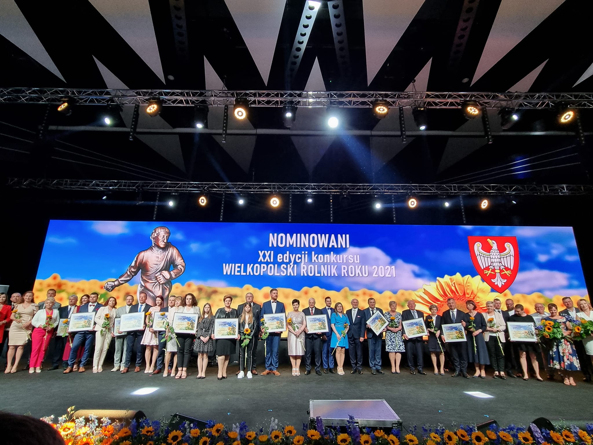 Grupa osób, stoją obok siebie. Część  osób trzyma piękne słoneczniki, pozostałe osoby mają w ręku dyplomy. Znajdują się  na dużej scenie, za ich plecami umieszczony jest ogromy telebim na którym wyświetlone jest pole ze słonecznikami